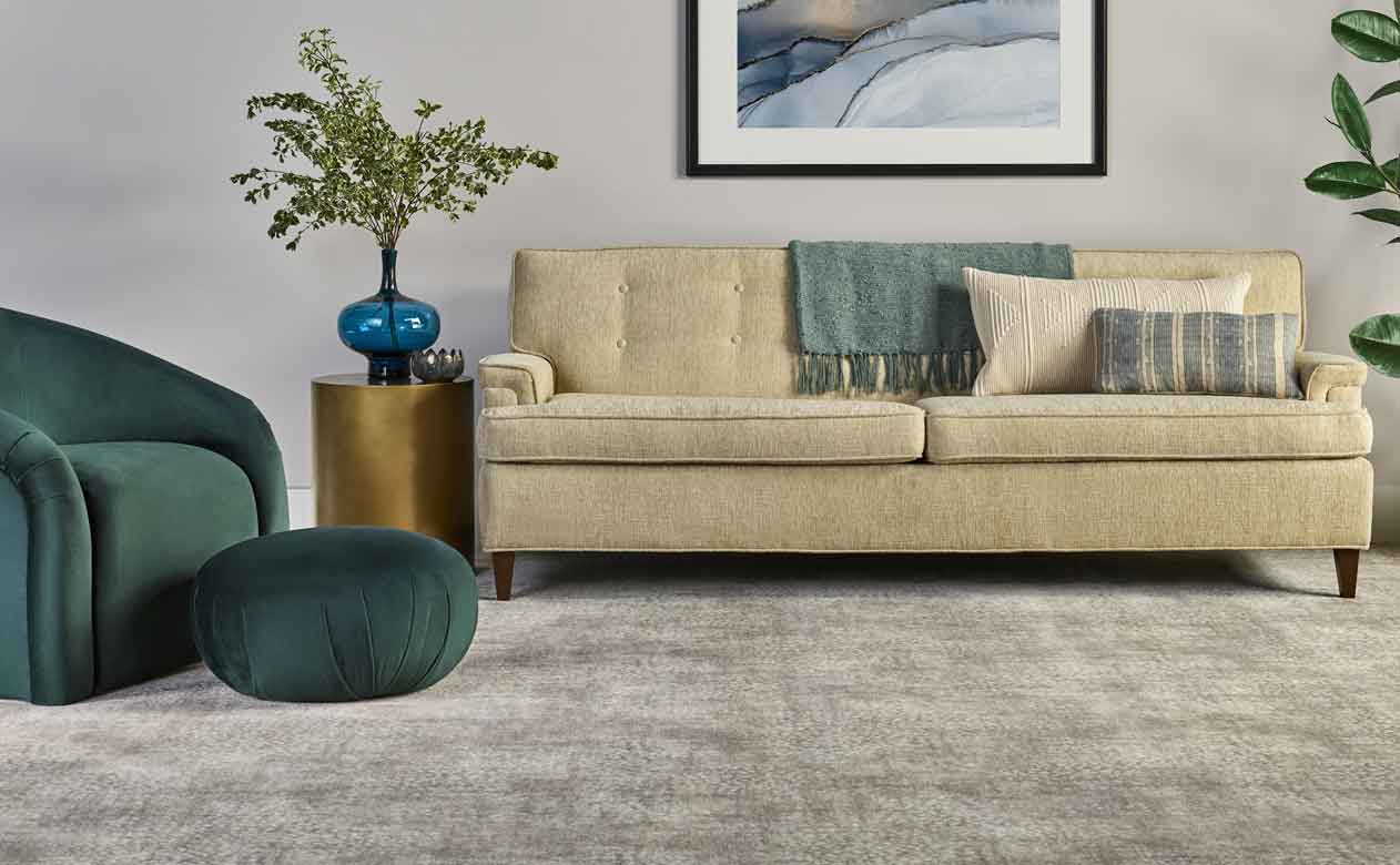 Plush greige patterened carpet in living room with beige couch, velvet green ottoman, and greenery 